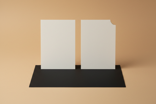 A minimal still life on the same warm beige background. Two identical off white cards sit side by side. One card is perfectly blank. The other has a subtle die cut corner or a distinctive edge profile, barely noticeable but intentional. Beneath them, a single sheet of heavy black paper is placed like a base, cleanly aligned. The scene communicates distinction through small decisions, not decoration. Soft diffused light, gentle shadow, no text, no logos.
