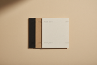 A restrained still life on a warm beige seamless background. A clean arrangement of three paper swatches: one matte black, one warm beige brown, one soft off white textured stock. Each swatch has a precise cut edge and a slight overlap, forming a quiet geometric composition. A faint blind emboss pattern is visible only through shadow and angle, suggesting brand codes without showing any logo. Soft diffused light, gentle shadow, minimal and tactile, no text.
